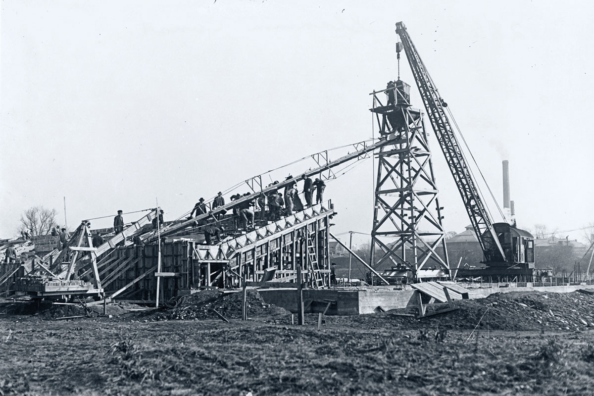 A team of workers construct one of the first sections of stands — only the wooden structure looks to have been put in and the men stand on top. The black and white photo includes a crane and wooden frame for a taller structure.