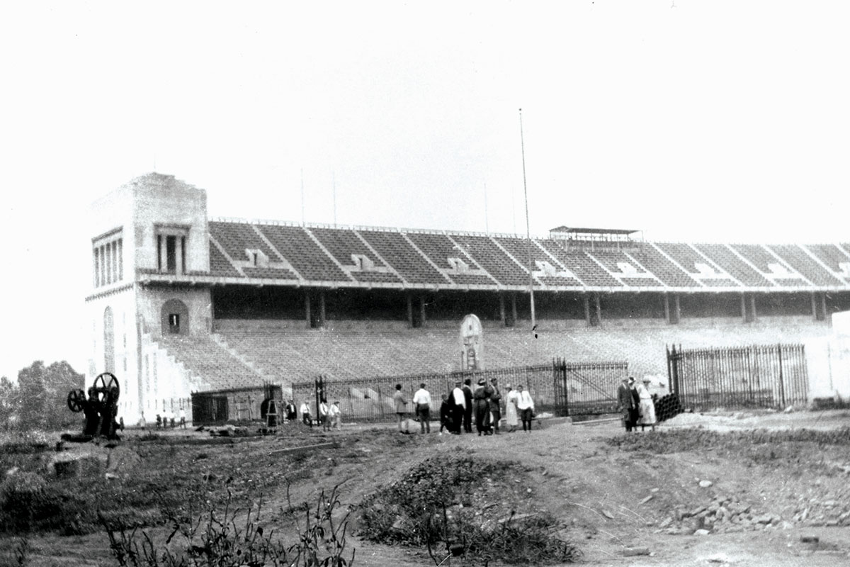 At a time when Ohio Stadium was almost completely constructed, more than a dozen people mill around outside the open end of the horseshoe. In the distance, one end of Ohio Stadium and its newly completed seating can be seen.