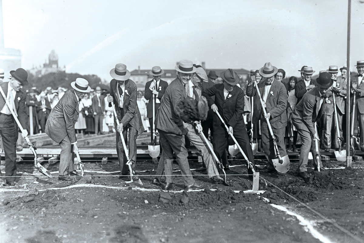 In an old black and white photograph, seven men in suits and hats are lined up and shoveling dirt where the stadium will be built.