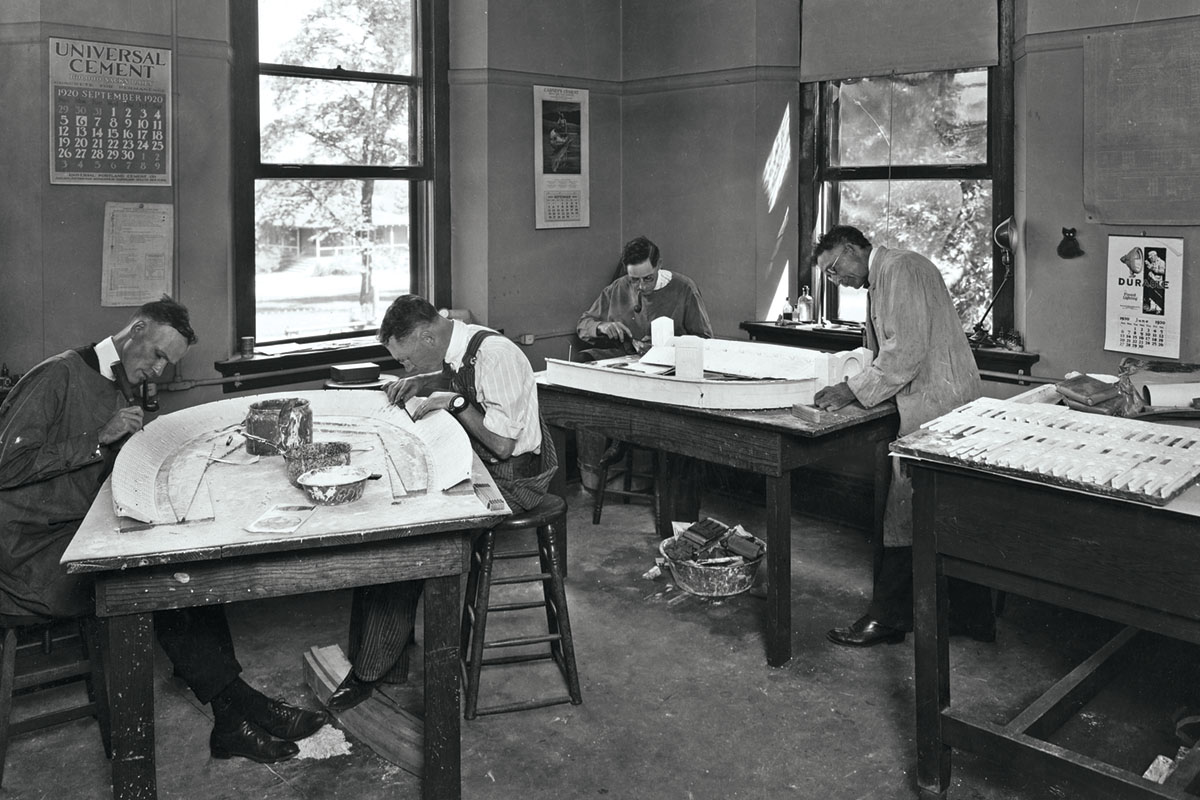 In an old black and white photograph, four men work at plain wooden tables building horseshoe-shaped stadium models.