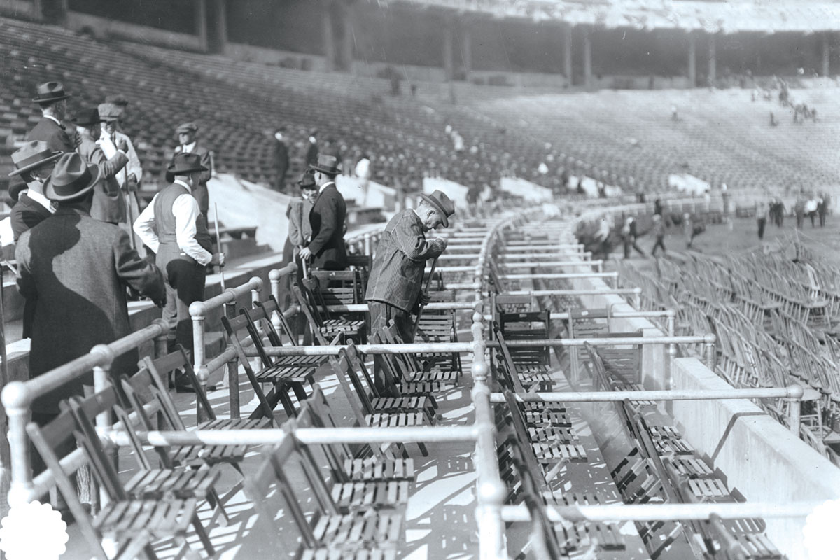 A historical photo shows men in suits and hats, each holding a broom, chatting amid the stands at Ohio Stadium. The concrete risers are filled with wooden folding chairs 