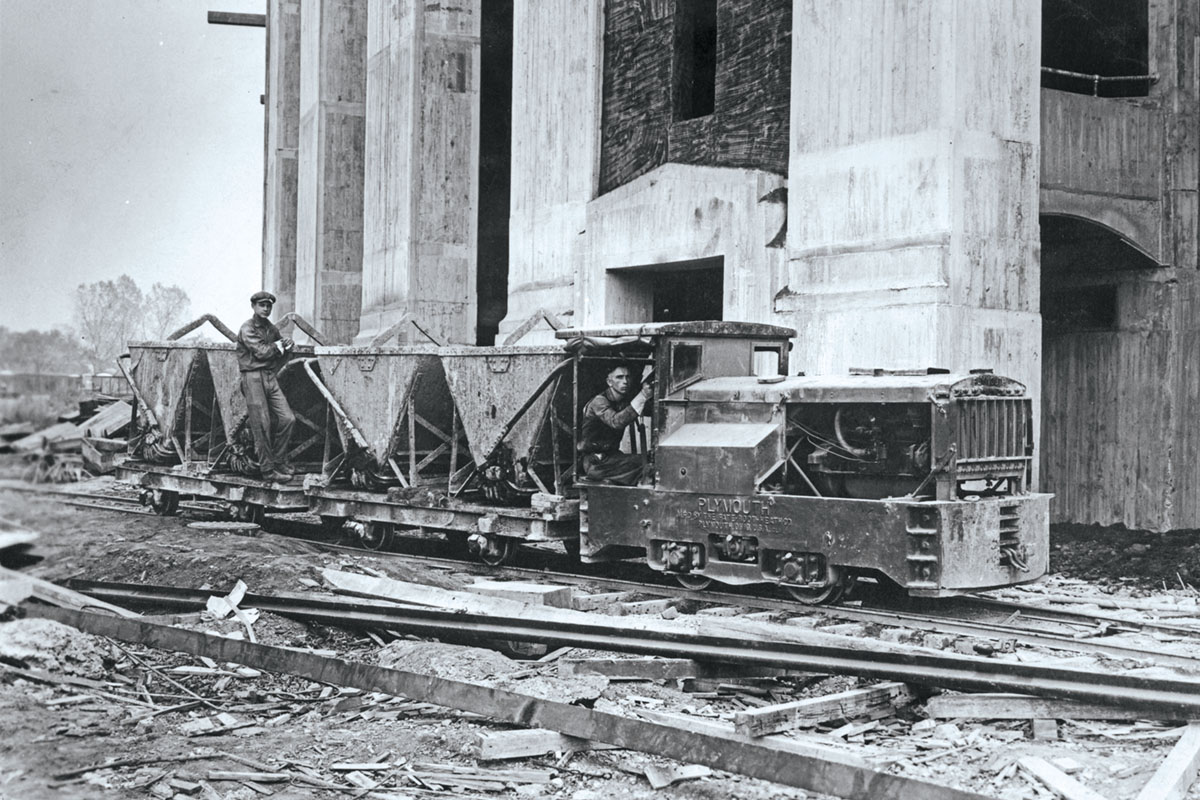 A small train engine pulls vats of cement on temporary tracks through the material-littered construction site.