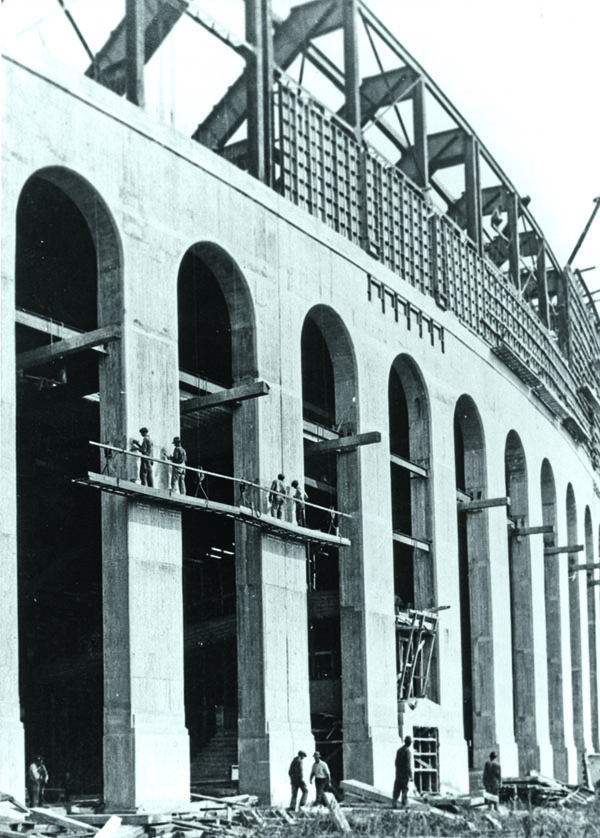 Concrete arches are in place in this vertical photo from the outside of the stadium, but the upper deck of stands is no more than lumber and steel. Men on a suspended scaffolding make adjustments to the front of the concrete, and men on the ground step over lumber and other construction materials.