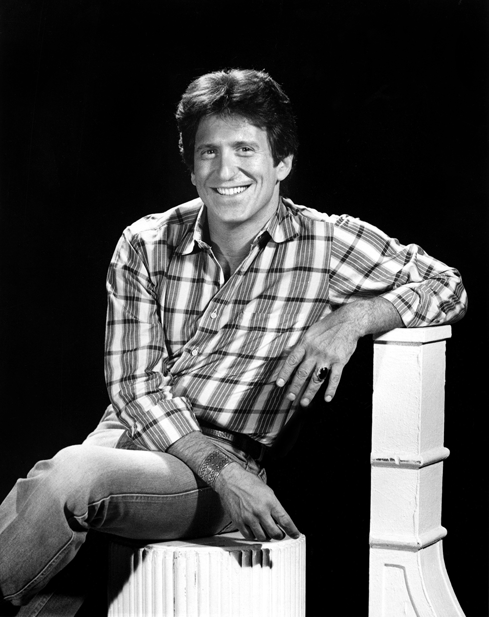 Michael Ochs smiles for a portrait as a younger man. A white man, he has thick hair, a big smile and wears a plaid shirt and jeans.