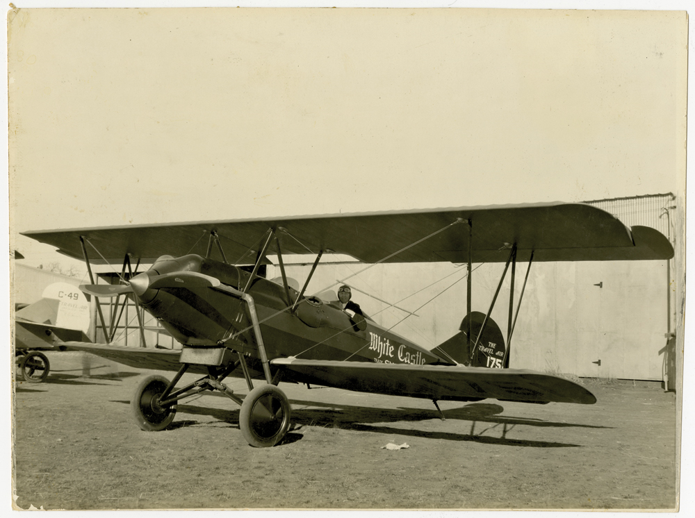 An old photo shows a pilot sitting in a biplane. The word "White Castle" are on the side.