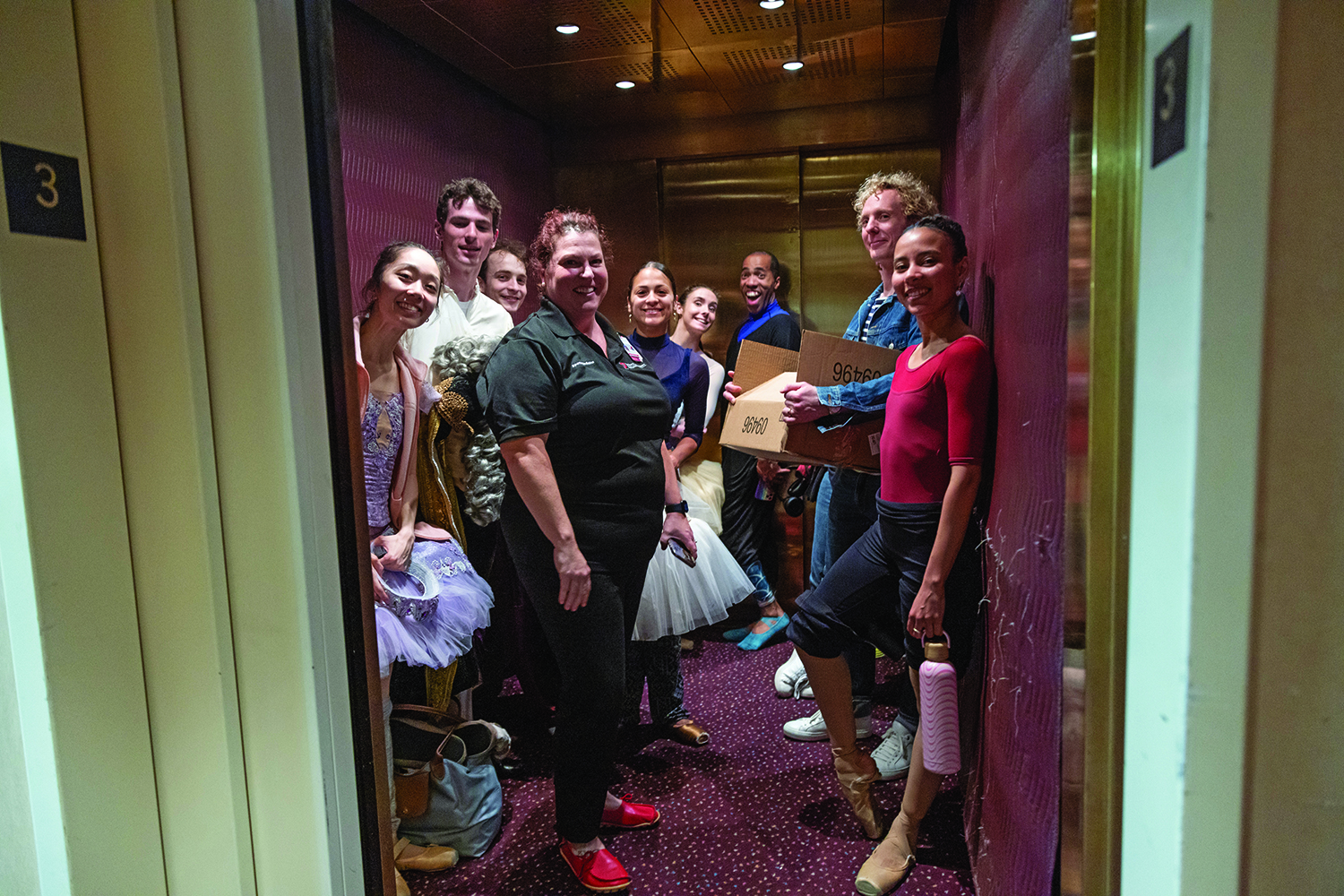 Dancers surround an athletic trainer inside a richly colored elevator. They all smile or mug for the camera.