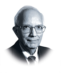 A man of Indian descent smiles while looking directly into the camera. He wears glasses and a suit and tie and has a close-lipped smile.