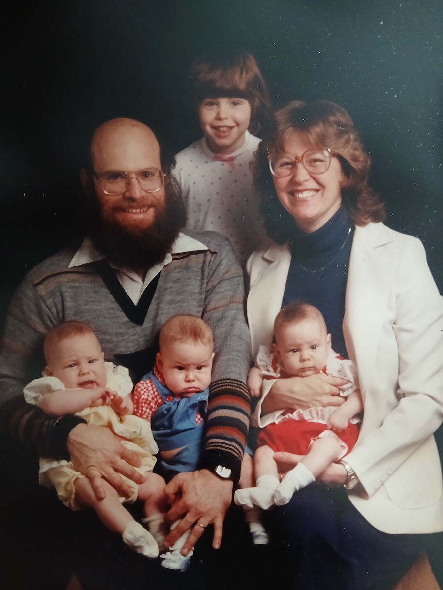 A professionally-taken family photo from the 1980s shows a mom and dad sitting with their oldest daughter centered behind them and their triplet daughters on their laps. The babies are probably not 1-year-old yet and none of them are happy to be there--though the rest of the family is smiling as if they're laughing.