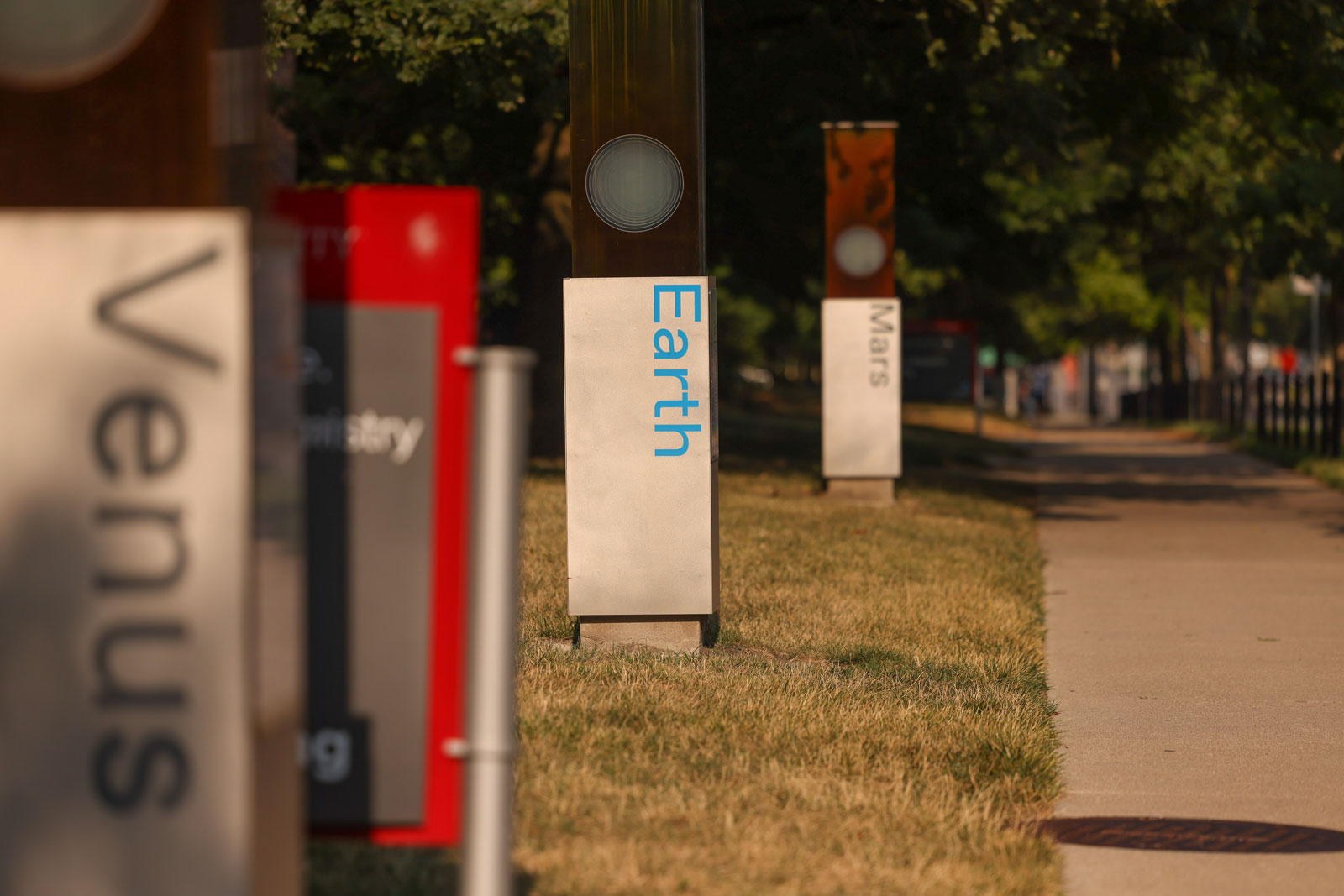 Outdoor stands naming planets—Venus, Earth, and Mars—are placed along a sidewalk in a grassy, tree lined area of campus. Atop each sign is a series of tall rectangular plates that, when viewed from the side, show a clear circle representing the size of the planet. The stands are placed at scale to the true distance they are from each other in the solar system.