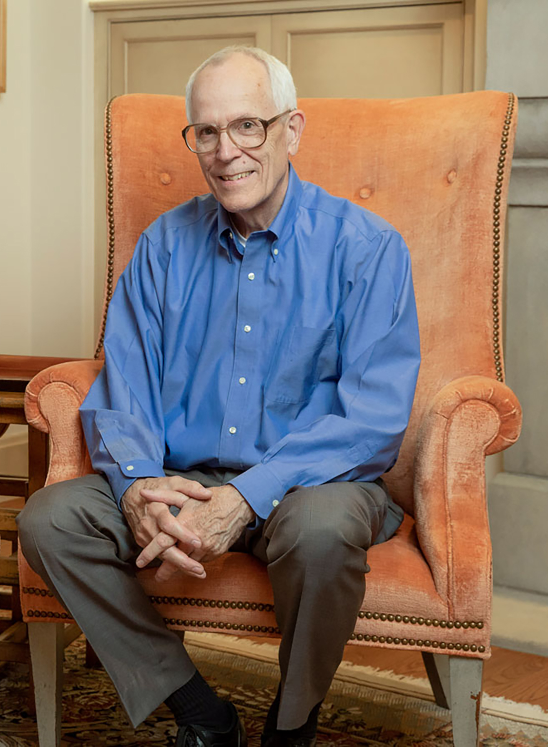Andy Ross smiles as he sits in a sophisticated upholstered armchair with hands loosely folded. He wears a button down, dress pants and glasses, and he looks like someone who would be interesting to have a conversation with.