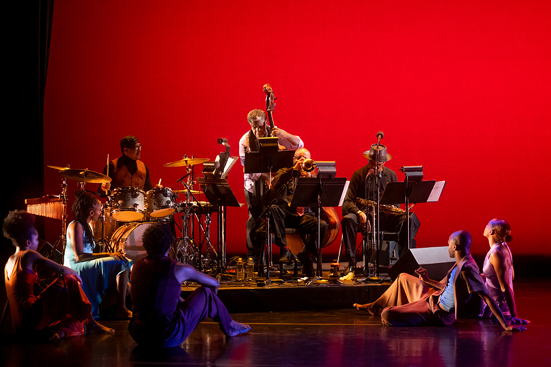 A four-piece band with, from left, a drummer, upright bass player, trumpet player and saxophone player, perform on stage. At this point in "In the Same Tongue," the dancers sit on the floor around the band, showing with their poses and expressions how engrossed they are in the music.
