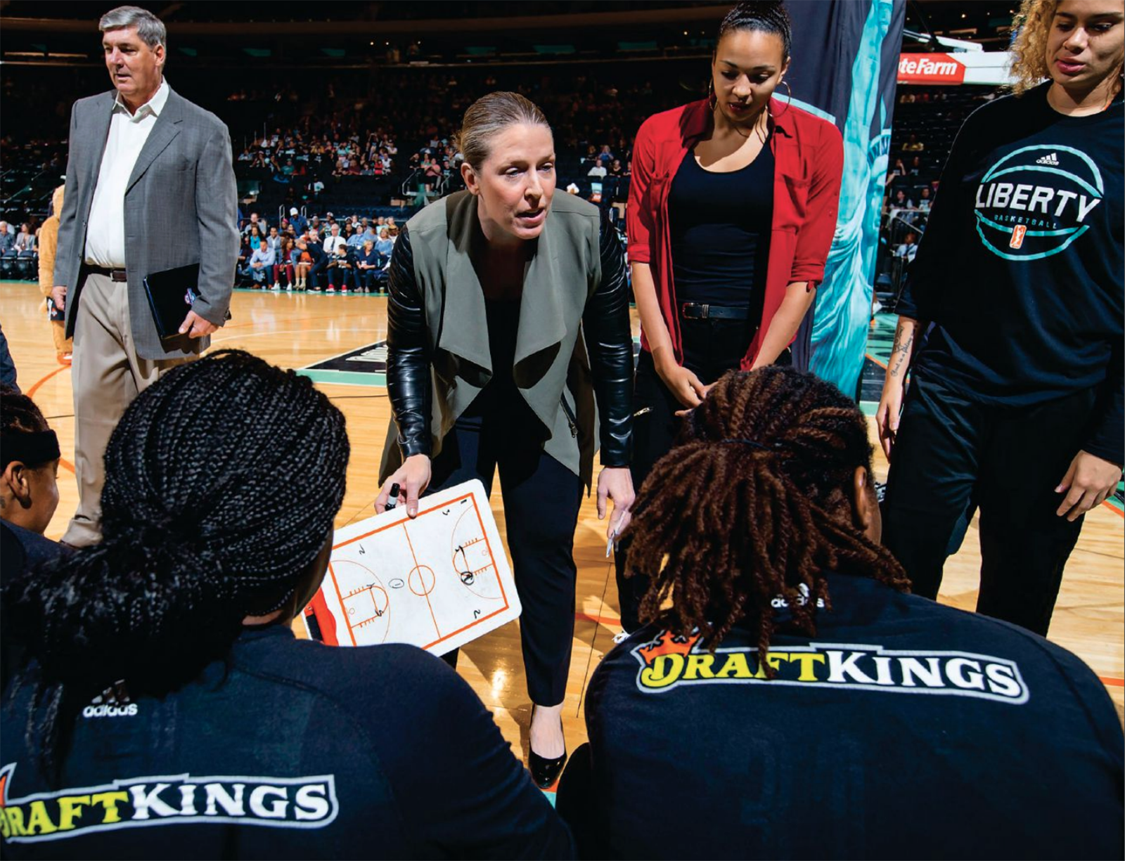 dressed in a stylish jacket with leather sleeves and her hair pulled back in a sleek ponytail, Katie Smith leans forward as she talks to women's basketball players inside an arena.