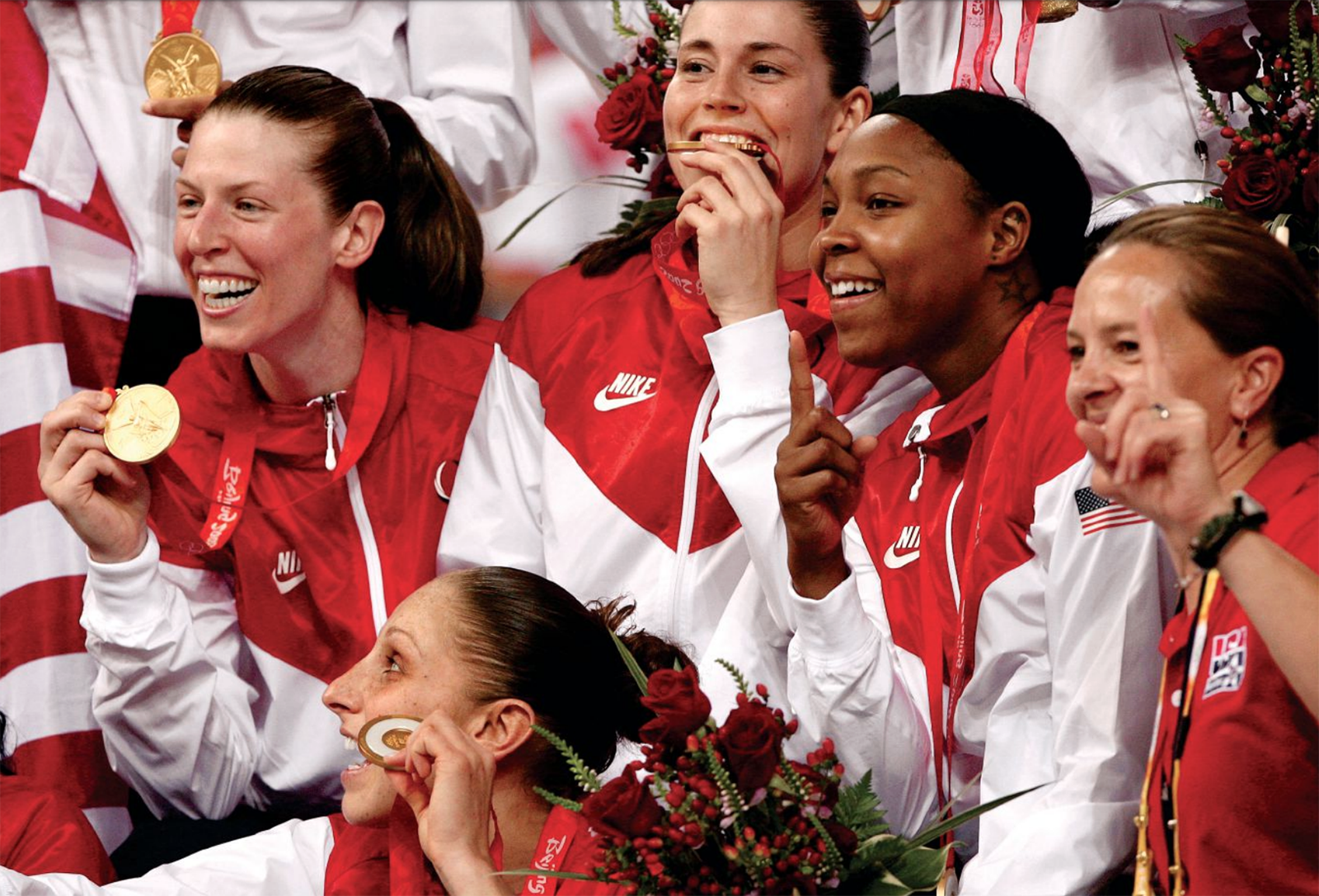 Katie Smith sits among teammates celebrating their Olympic Gold Medals, which they grin as they show off.