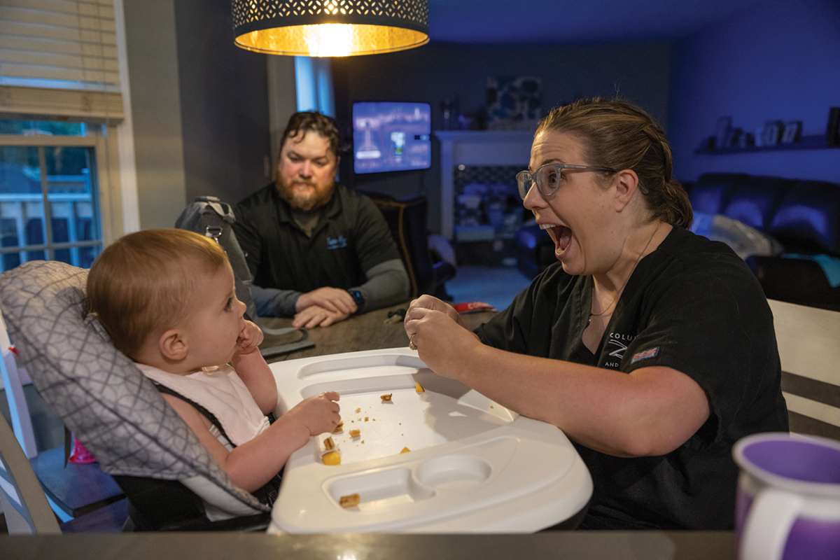 A mom gives her baby a massive grin as she feeds her in a high chair, while the dad sits at the table and watches.