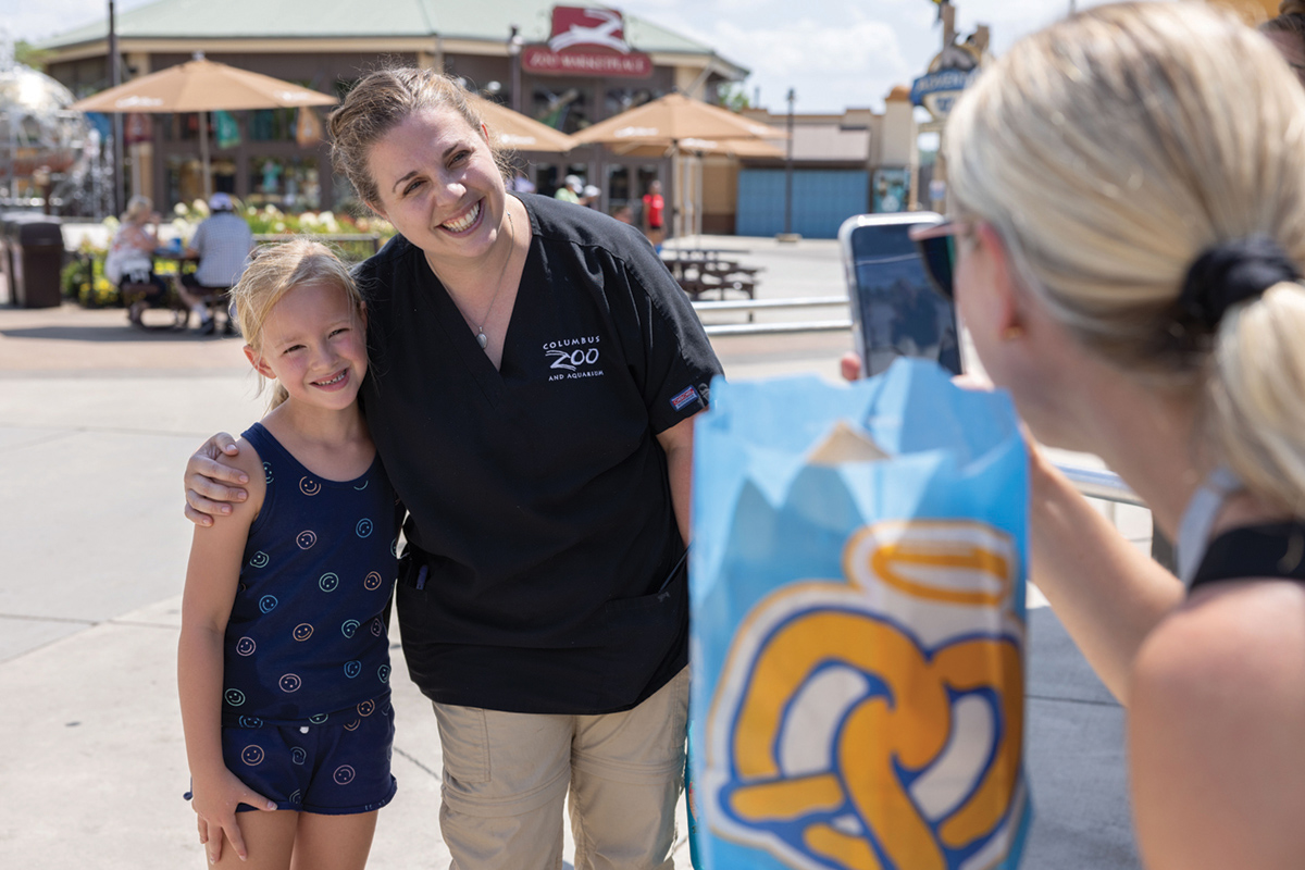 Outside at the Columbus zoo, a woman wearing a zoo vet shirt leans to wrap her arm around a blonde girl. Both smile as the kid’s mom takes a photo with her cellphone.