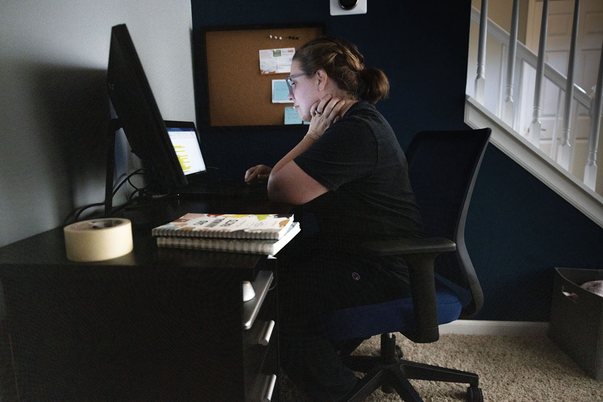 A woman sits at a desk in a dim room working on two monitors. She looks engaged yet tired.