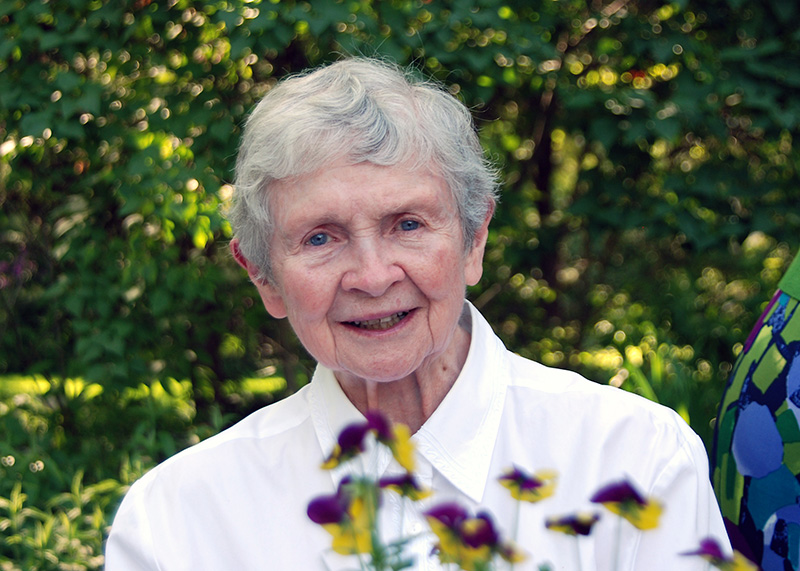 Sister Mary Faith Dargan, an older white lady, smiles as she poses for a portrait outdoors. She wears a plain white button-down that makes her blue eyes and smile stand out