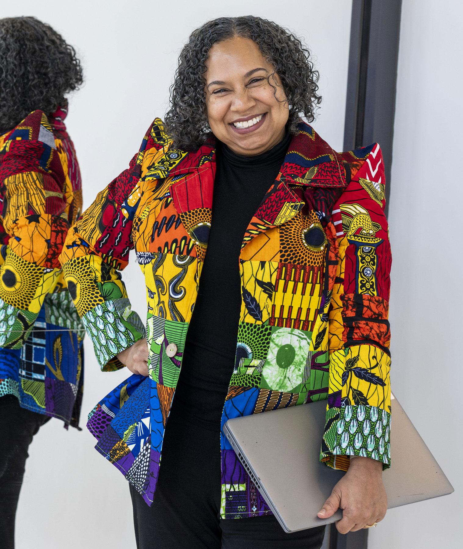 A Black woman grins as she holds a closed laptop and stands in front of a large mirror. IT reflects the back of her curly hair and patterned, stylish jacket. The hand not holding her laptop is on her hip. She comes across as bright, sunny and well-styled.