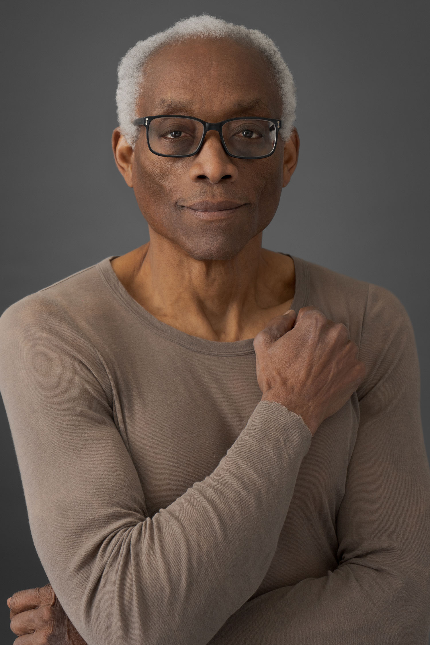 Bill T. Jones smiles slightly, appearing calm and composed while posing for this photo. He is an African-American man with white hair and glasses, and a bearing you’d expect from a dancer.