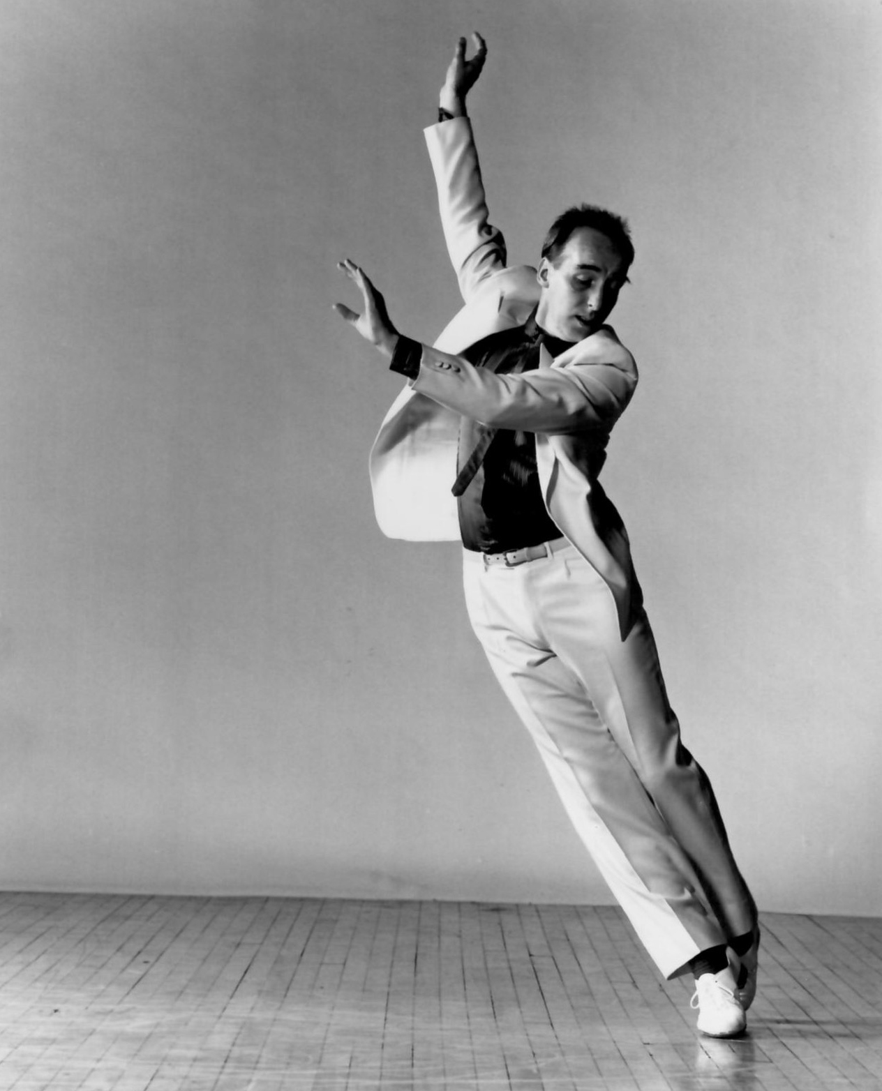 Fred Strickler tap dances with his full body slanted in a diagonal. He reaches up and out and looks down toward the stage.