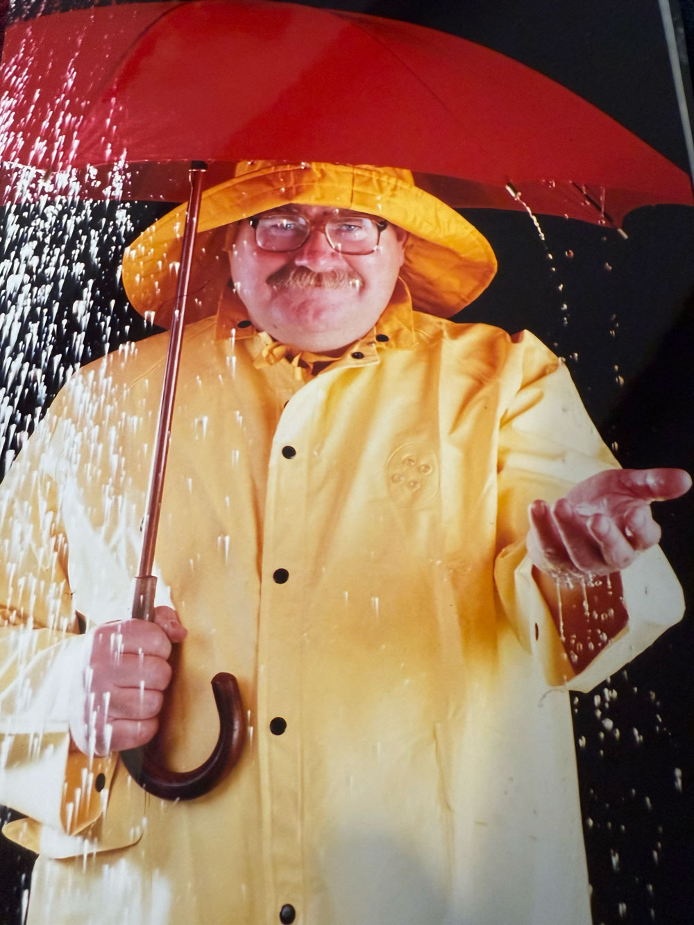 A person wearing a yellow raincoat and holding a red umbrella stands in falling water, with one hand extended outward to catch the rain. He is John Switzer and he has a mischievous smile, big glasses and a big mustache that hint at his humor