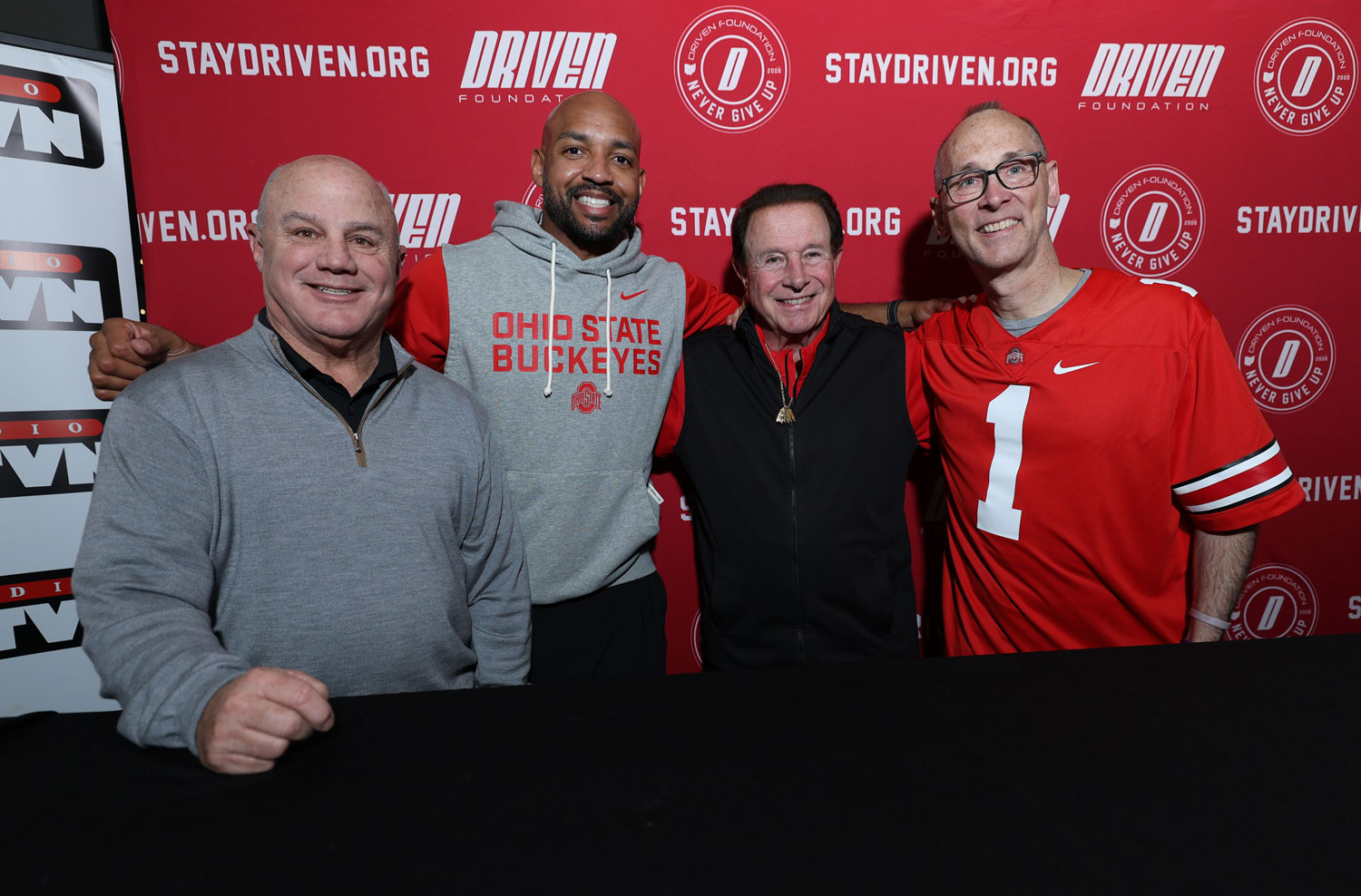 Four men stand with their arms around each other and smile.