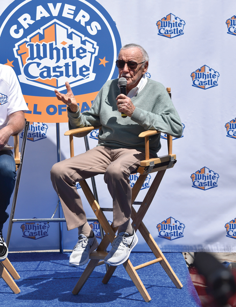 Stan Lee sits in a director's chair and talks into a microphone in front of a White Castle backdrop