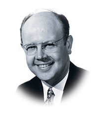 A white man with glasses grins as he poses in his suit and tie.