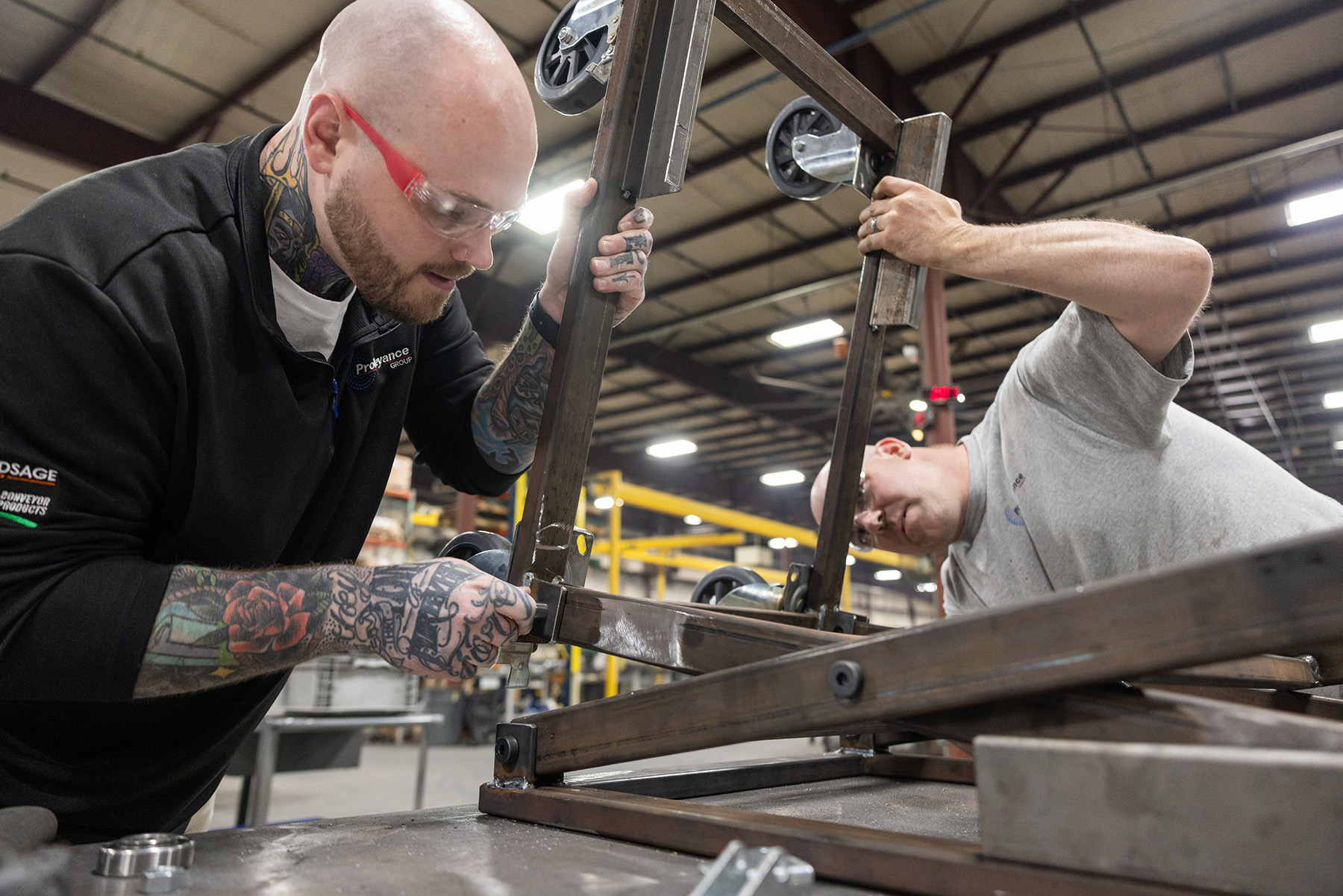 Two men work on assembling part of the metal system that will be a conveyor belt. Both wear safety glasses and have shaved heads. They look focused, and like they work well together. 