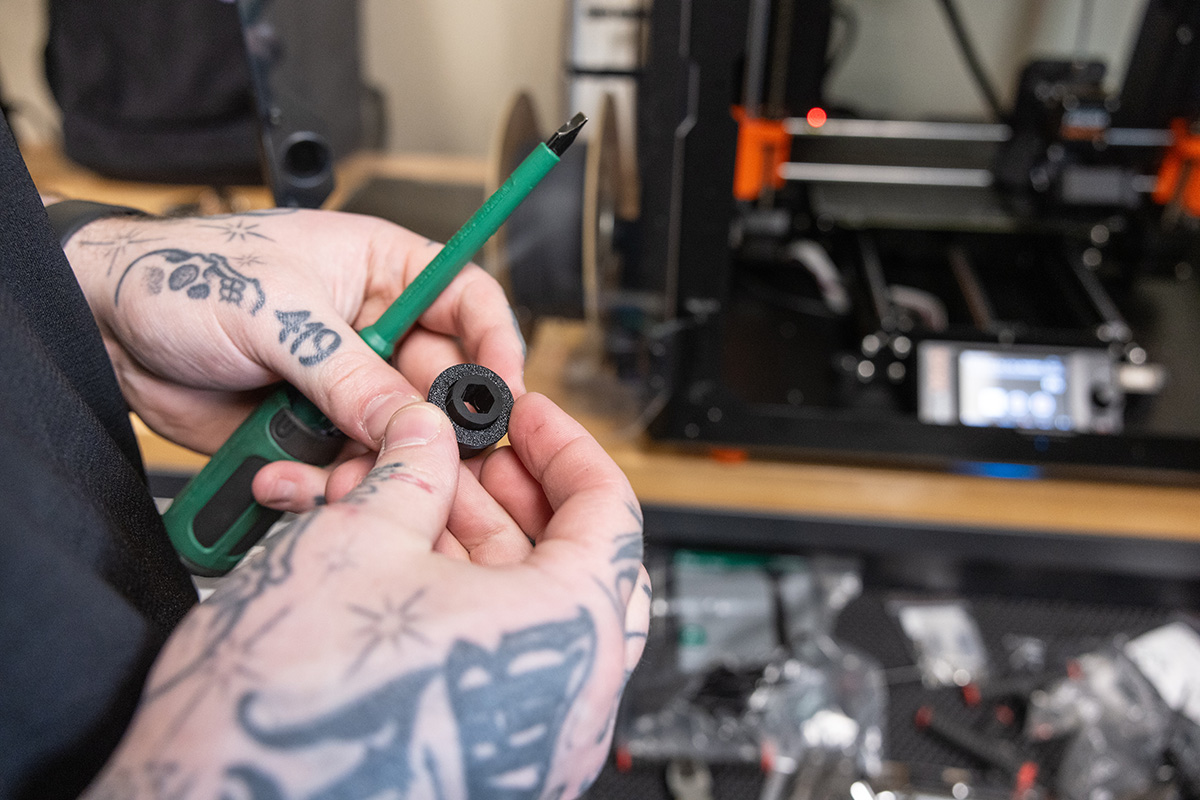 A photo shows Zack's hands as he holds a small round piece with a hexagon hole in the middle and a slim screwdriver. His hands have tattoos of letters, star bursts, part of a skull and the numbers 419.