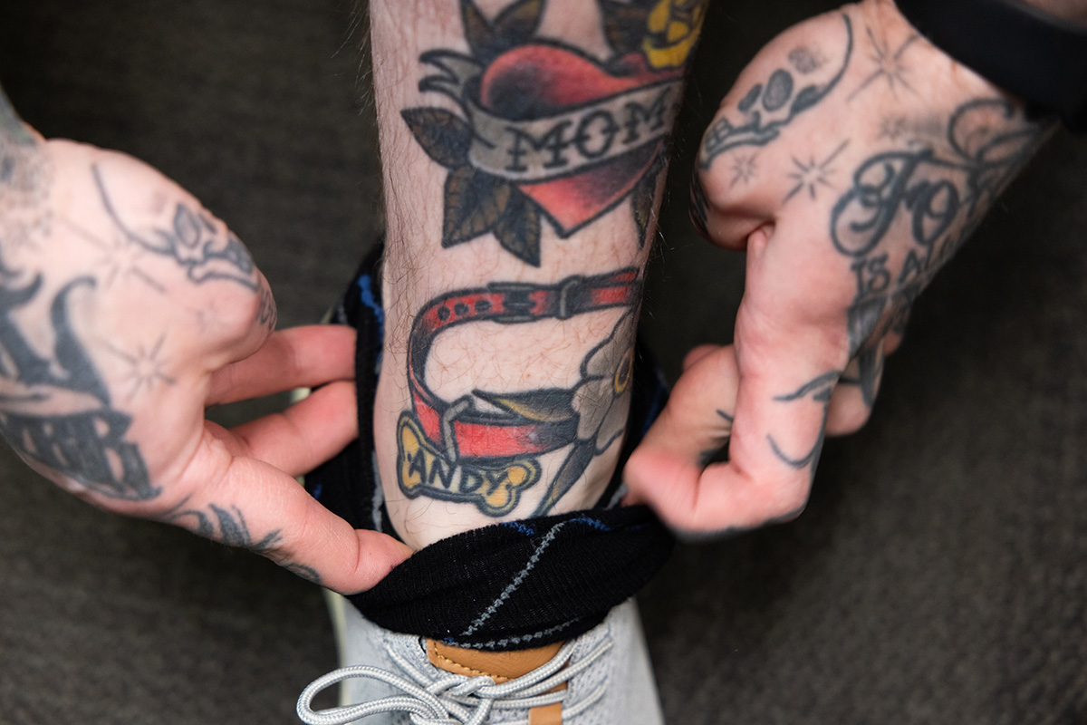 Zack pushes down his sock to show tattooes on his leg. There is a heart with a "mom" banner across it and a dog collar with a tag that says "Andy," as well as flowers and leaves.