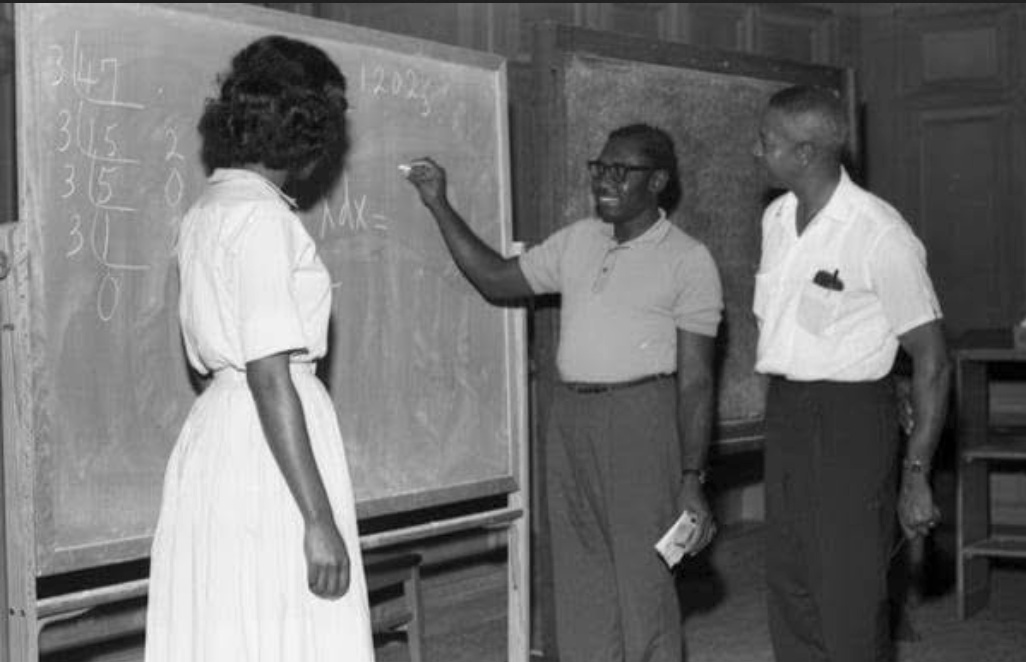 In an old black and white photo, a young Ossiefield Anderson works a math problem with chalk on a blackboard. An older man and a woman wearing a 1960s-style dress watch.
