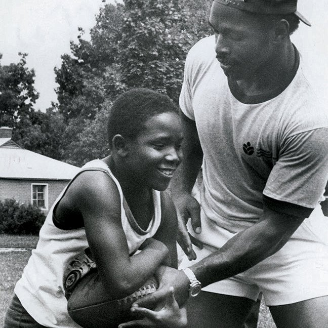 A young black boy tucks a football against his chest as he plays in the yard of a house with a man who is probably his dad. The photo is an old black-and-white one and the dad is reaching for the football. The boy, who is Archie Griffin, looks like he’s having fun.