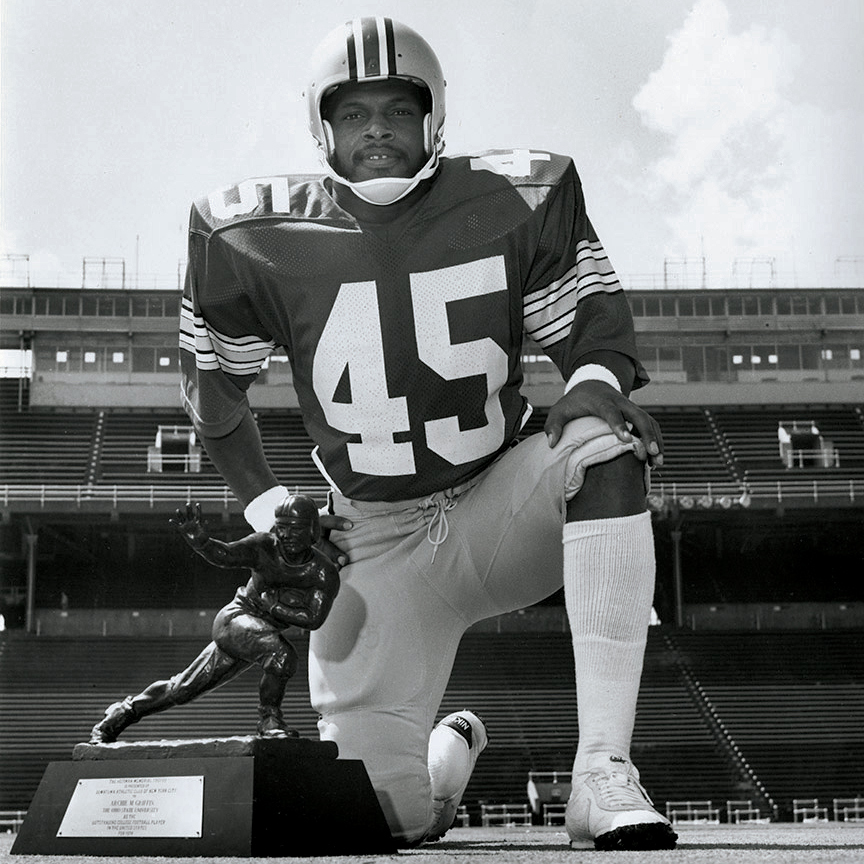 A college-age Archie Griffin kneels on The ‘Shoe’s otherwise empty football field with a Heisman trophy in front of him. He’s on one knee with his other leg forward and that foot on the ground and he’s wearing his No. 45 football uniform and helmet. His expression is low-key proud.