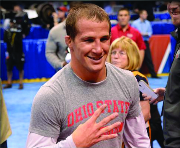 Wrestler Logan Stieber grins as he holds up four fingers on the sidelines of a meet