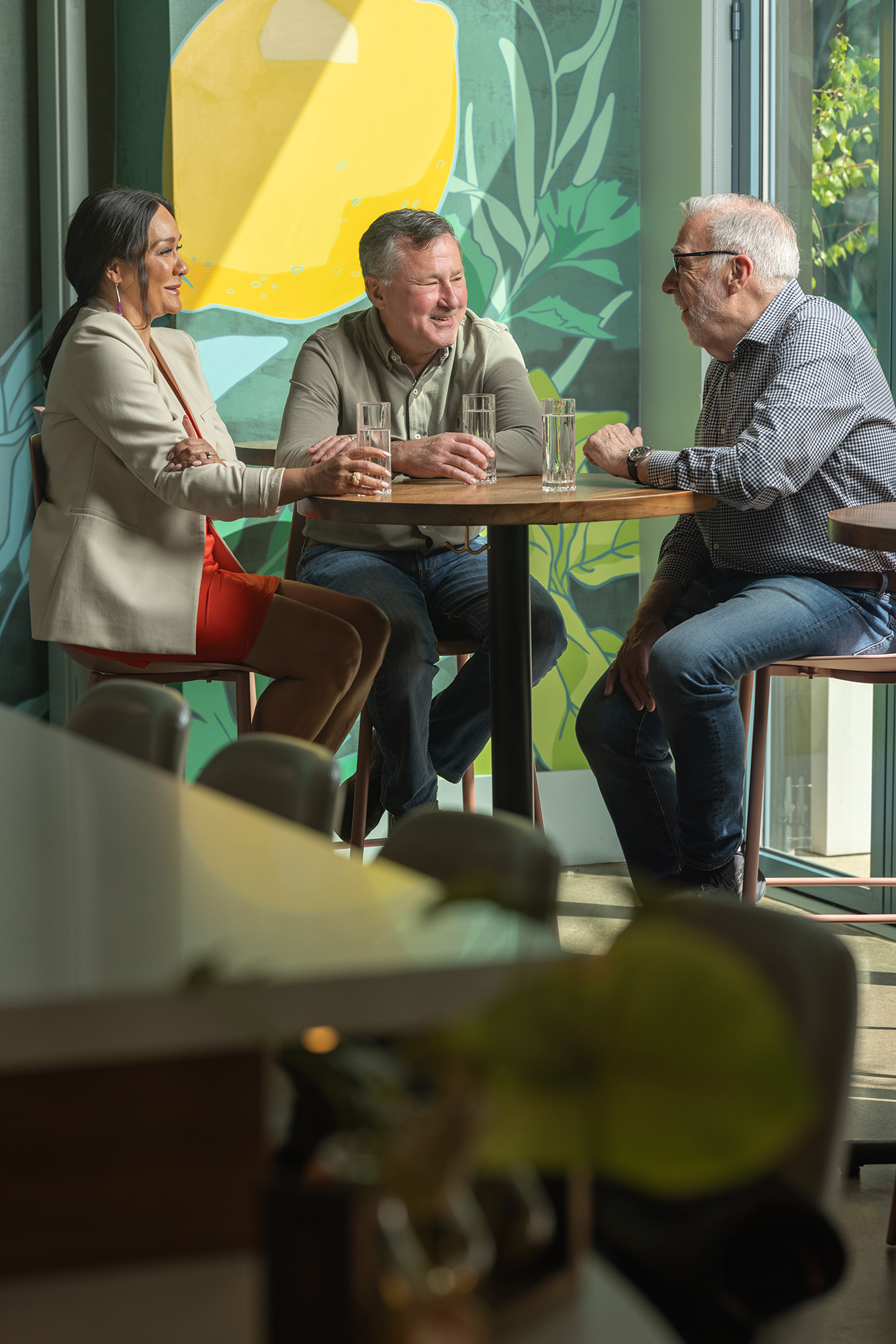 Michelle Brandt, Jeff Edwards and Duff Lindsay sit at a hightop table chatting. Behind them is a bold, colorful painting of lemons and greenery