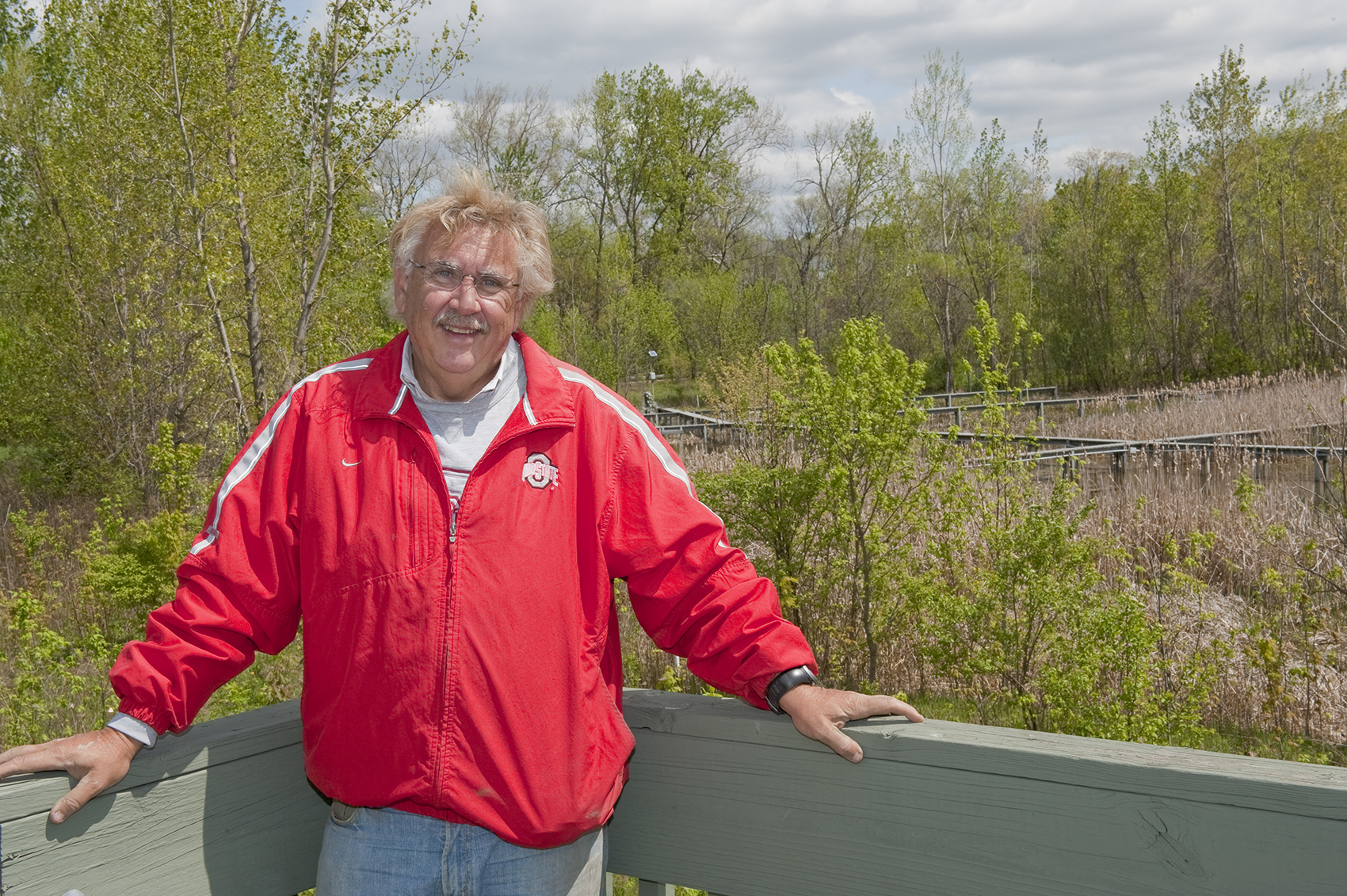 This portrait shows Bill Mitsch, a middle-age white man with a friendly smile. He's posing in front of the Olentangy River Wetland Research Park, which has raised wooden walkways over wetlands grasses inside a ring of trees.