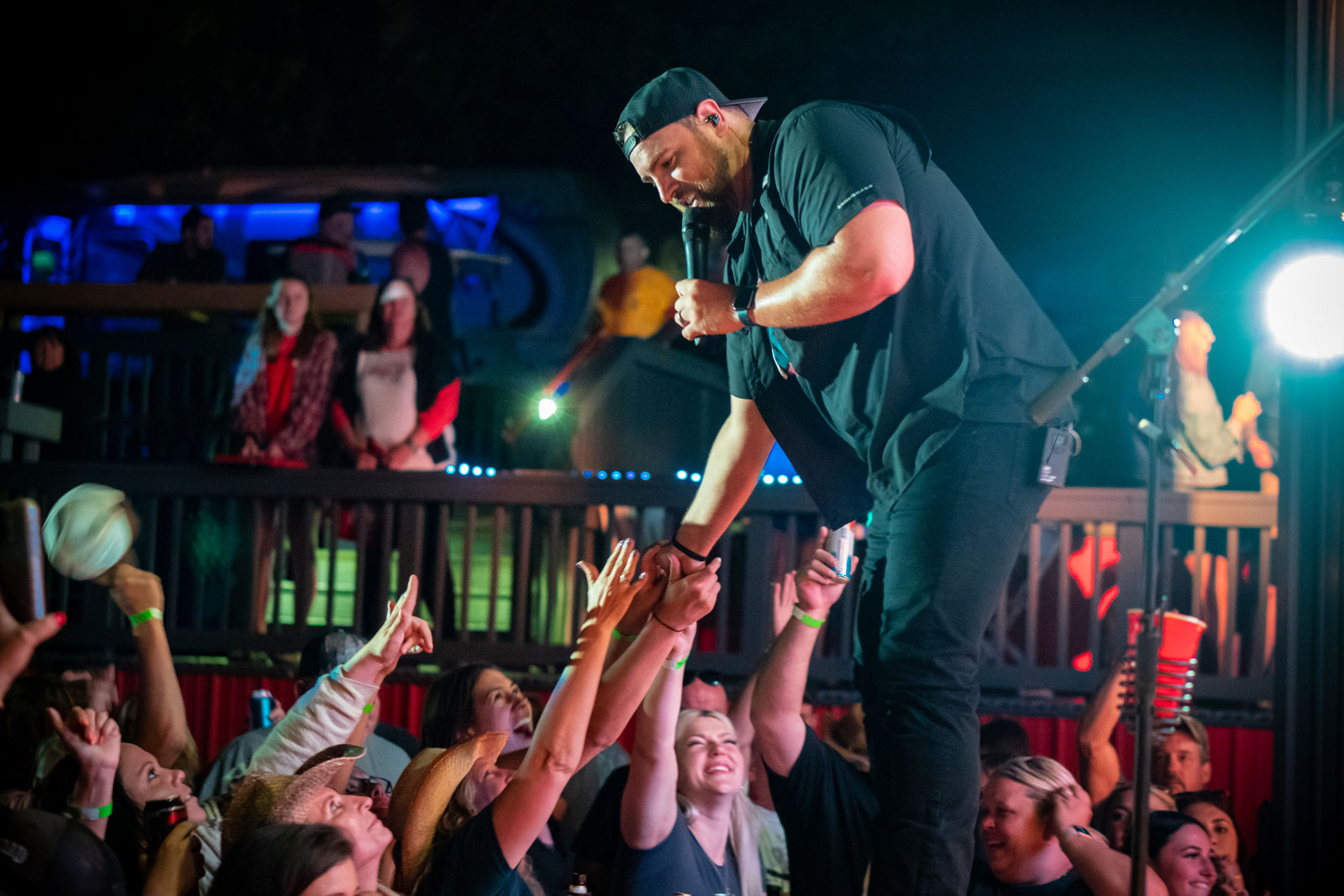 A man holding a mic on stage reaches down to clasp hands with adoring audience members. He wears all black and a backwards baseball cap.