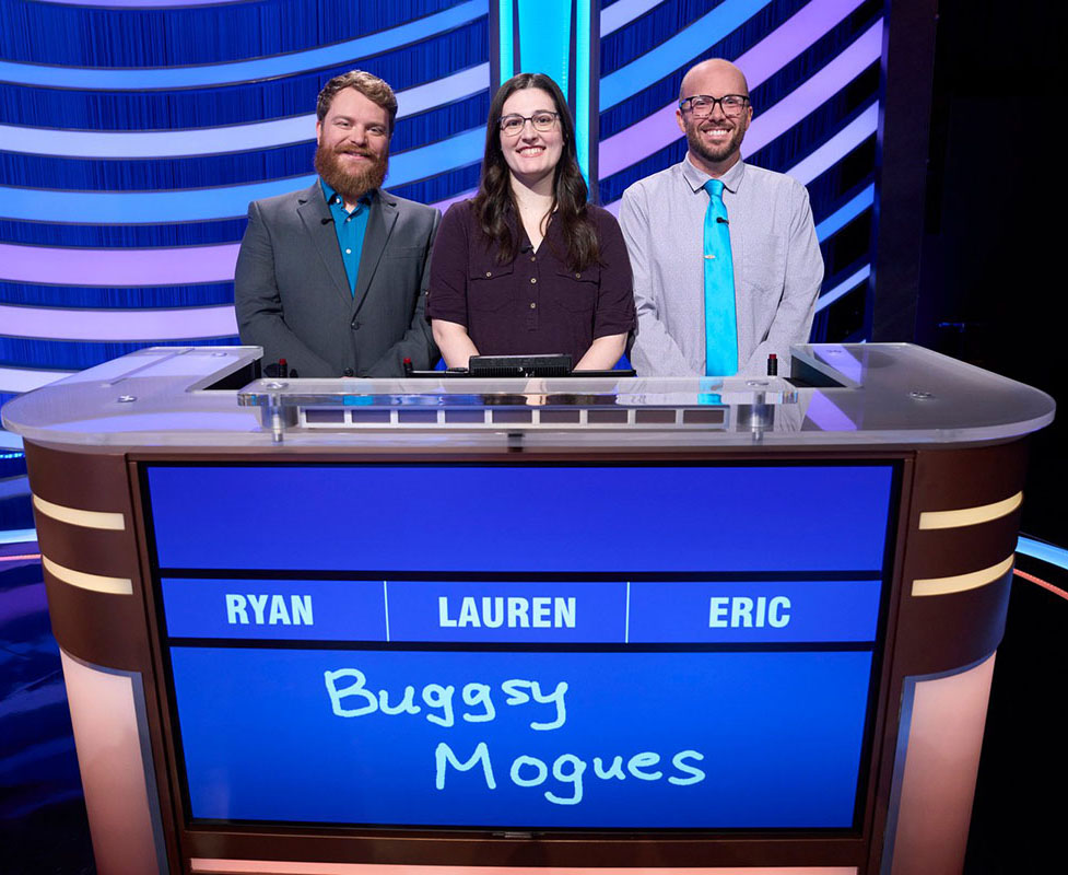 Two men stand on the sides of a woman on the Jeopardy stage. They're all smiling nicely and standing up straight.