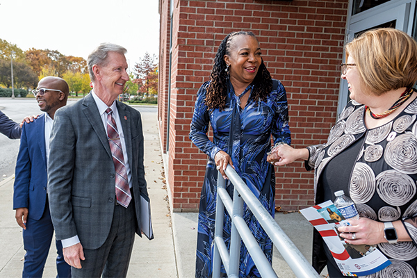 Standing outside a red brick building, President Carter smiles as noted alumna Stephanie Hightower and University Leader Stacy Raustauskas clasp hands and they talk.