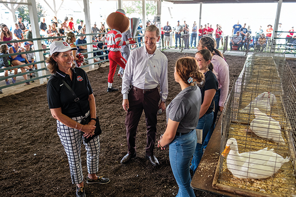 At a county fair, First Lady Lynda Carter and President Carter talk with three teens standing in front of cages with orange-billed white ducks. Brustus Buckeye is in the background in front of spectators sitting in stands. 