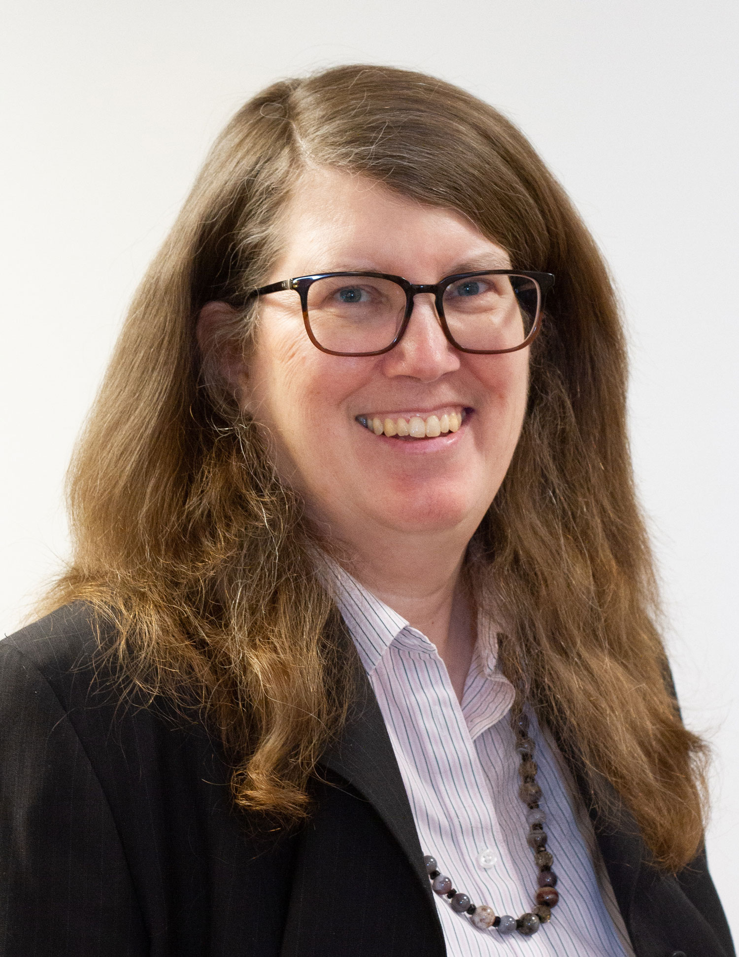 With long wavy hair and wearing glasses, a dark blazer and a beaded necklace, Ann Christy grins in a genuine way in this headshot.