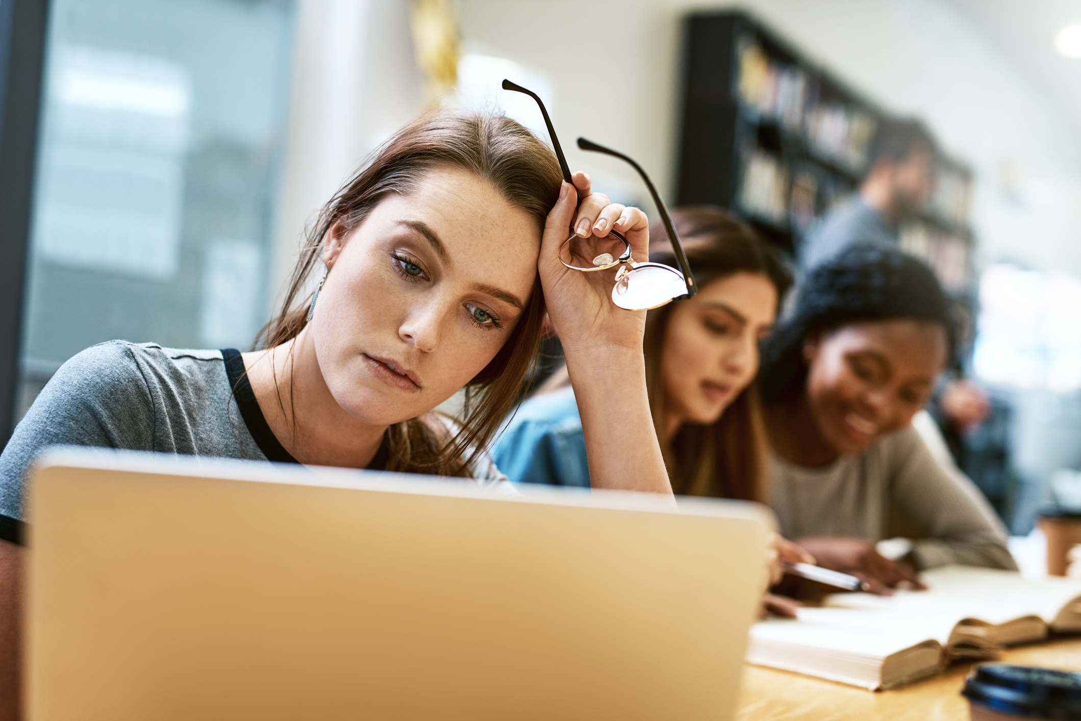 A stressed-out college student has her glasses off but looks at her computer screen while others in the background happily study.