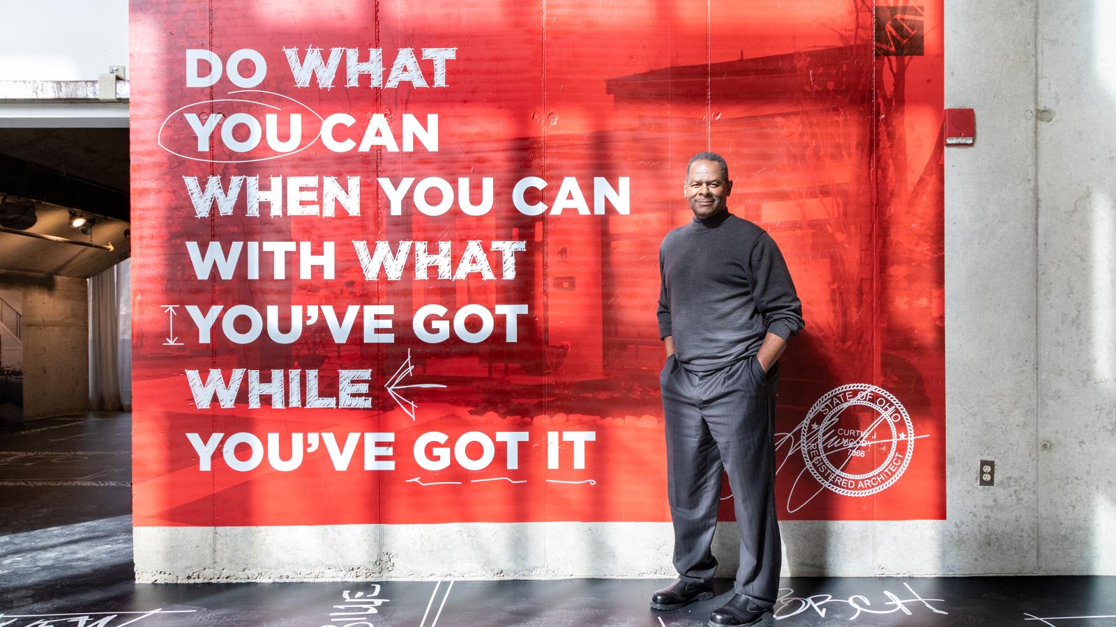 Curt Moody, a confident looking Black man,  casually poses for this photo in front of a painted concrete wall that says "Do what you can when you can with what you've got while you've got it.