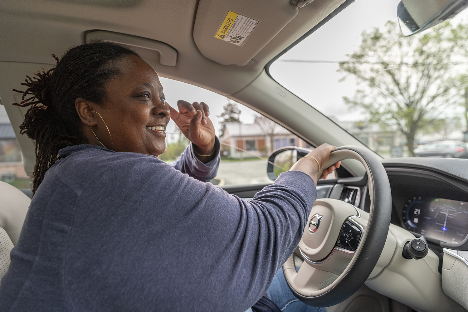 Daisy, a Black woman with her braided hair toed back, smiles as she drives her car to her next location. This photo was taken from inside, in the passenger's seat.