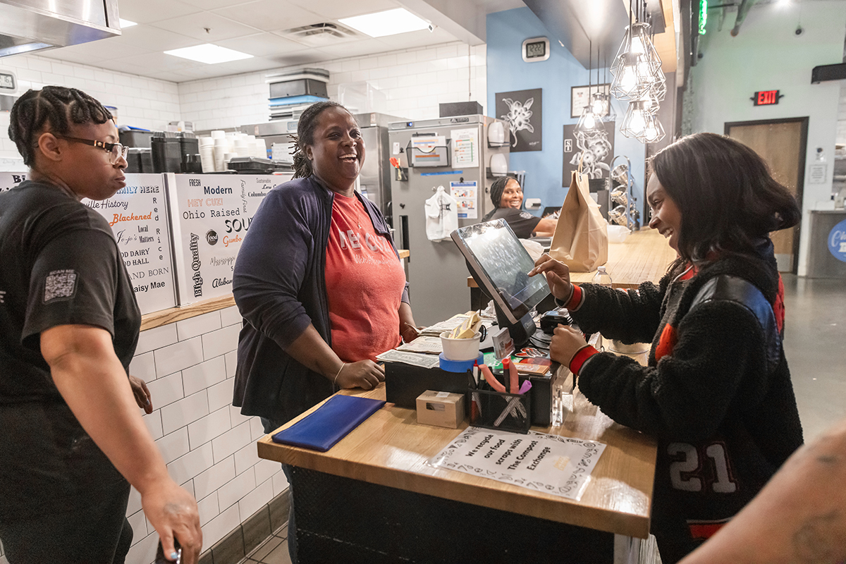 Daisy laughs as she greets a customer at the counter of her restaurant. Two employees are there with her.