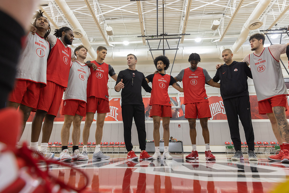 The men's basketball team stands in a semi-circle on the practice court with arms around one another's shoulders. Jake Diebler stands near the center and the players all look at him.