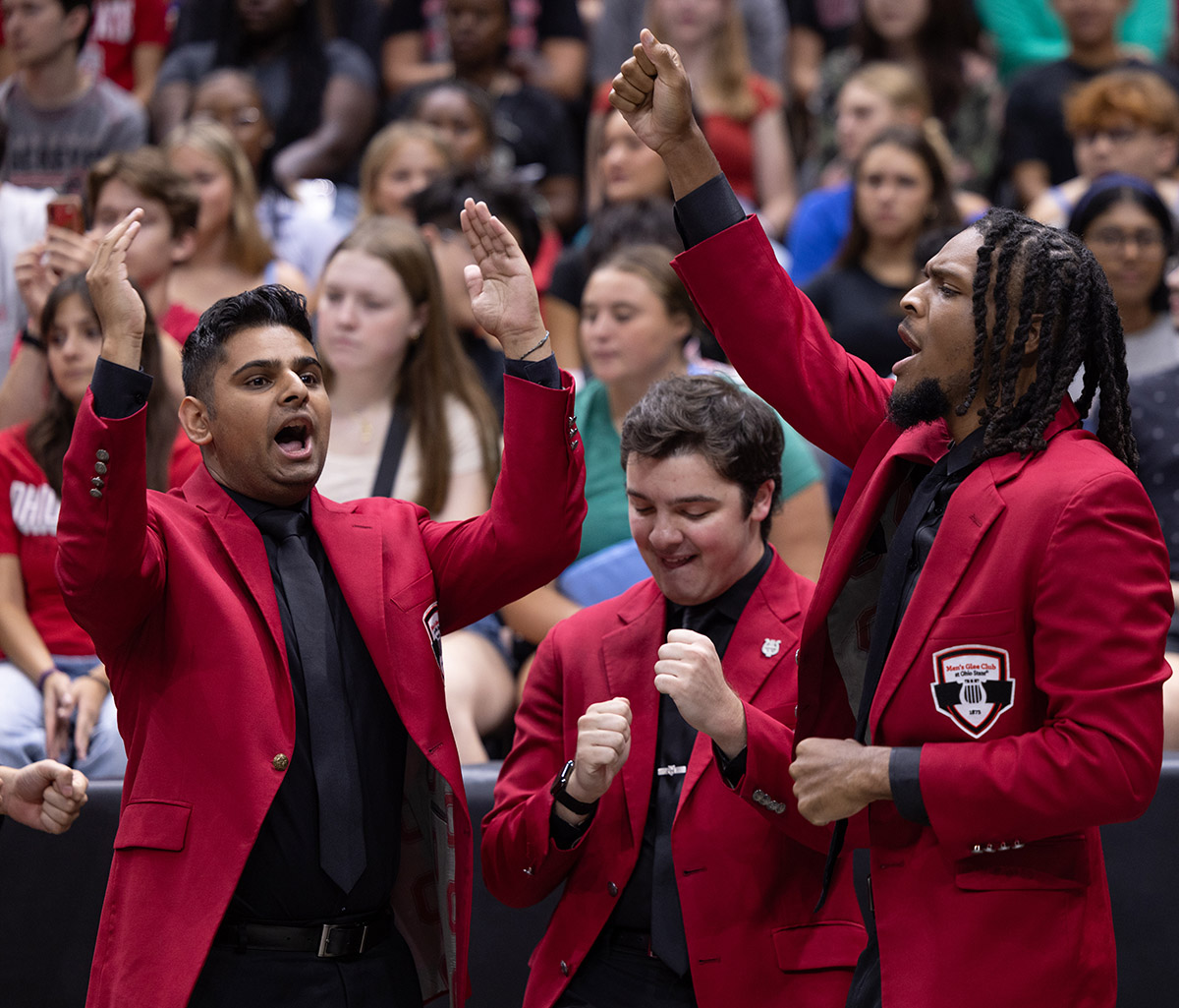 Three members of the men's glee club, all wearing scarlet sports coats over black button-downs and ties, dramatically sing in front of a crowd.