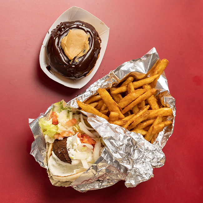 A tinfoil-wrapped gyro, with a falafel ball, lettuce, chopped tomatoes and tzatziki sauce, sits on top of a mound of French fries, each a little slimmer than a person’s pinky finger. Next the food is a doughnut topped with a thick coat of chocolate with a mound of peanut butter in the middle.