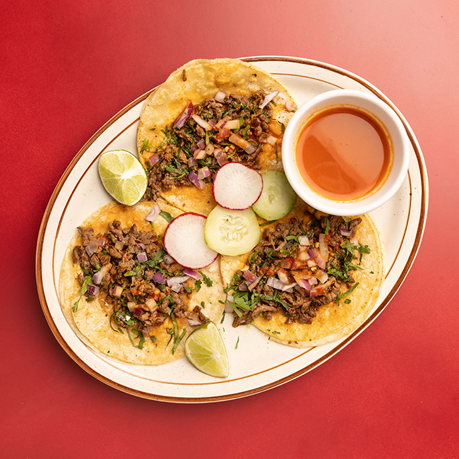 Three tortillas on a plate hold chopped-up taco meat. They’re accented by slices of radish, cucumber and lime. A small bowl of hot sauce accompanies the food.